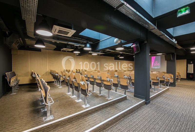 Salle de conférence moderne avec rangées de sièges en bois, éclairage au plafond et écran de présentation allumé.