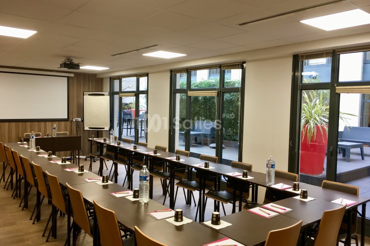 Salle de réunion lumineuse avec tables disposées en U, chaises, écran de projection et vue sur une terrasse extérieure.