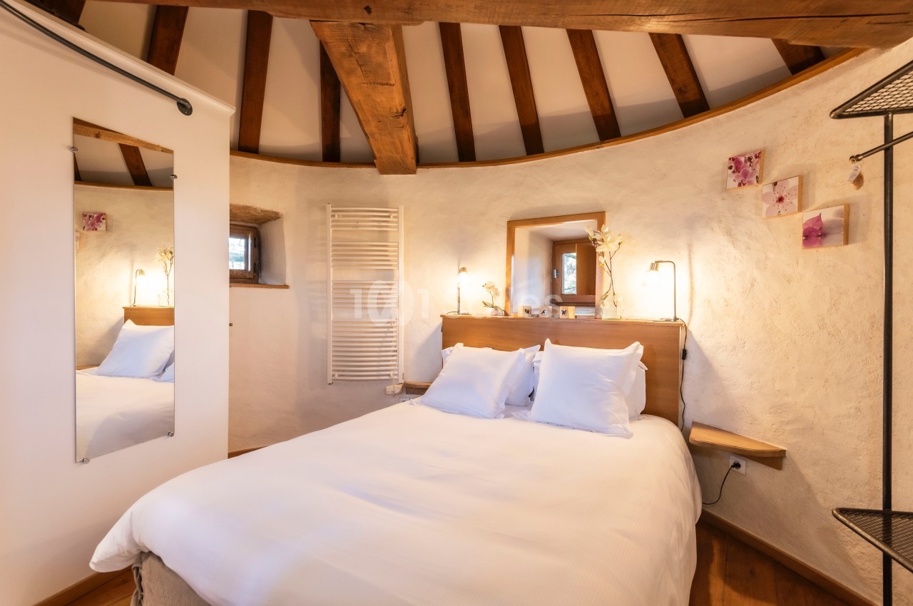 Chambre lumineuse avec lit double, murs en pierre blanche, poutres apparentes et décoration minimaliste.