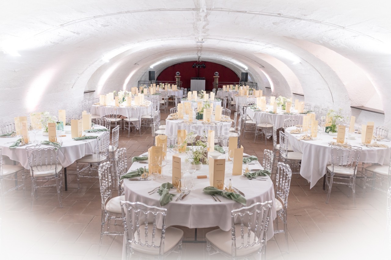 Salle voûtée décorée pour un banquet, avec tables rondes dressées, nappes blanches et chaises transparentes.