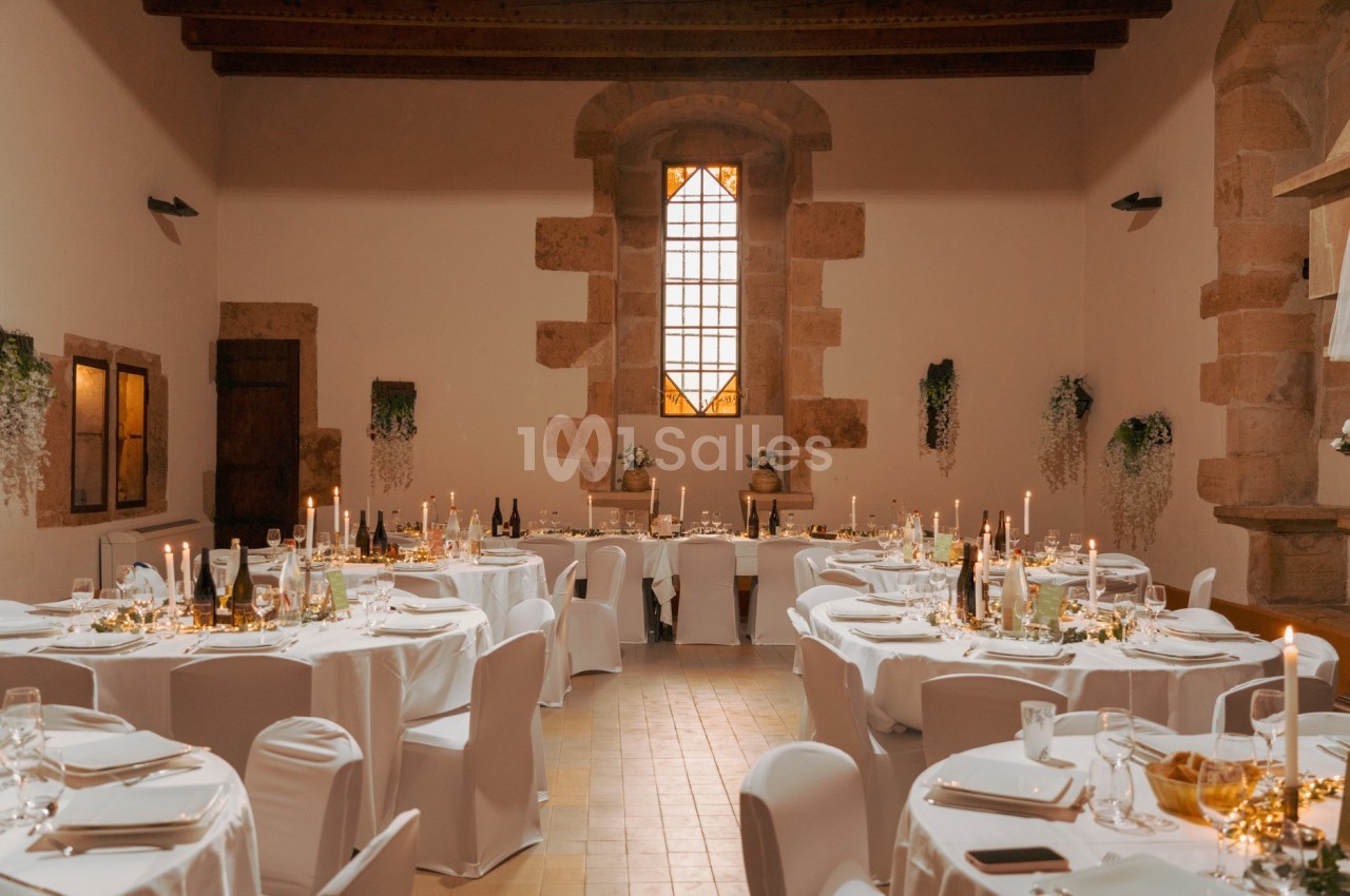 Salle de réception décorée avec des tables rondes, nappes blanches, chandeliers et lumière naturelle provenant d'une grande…