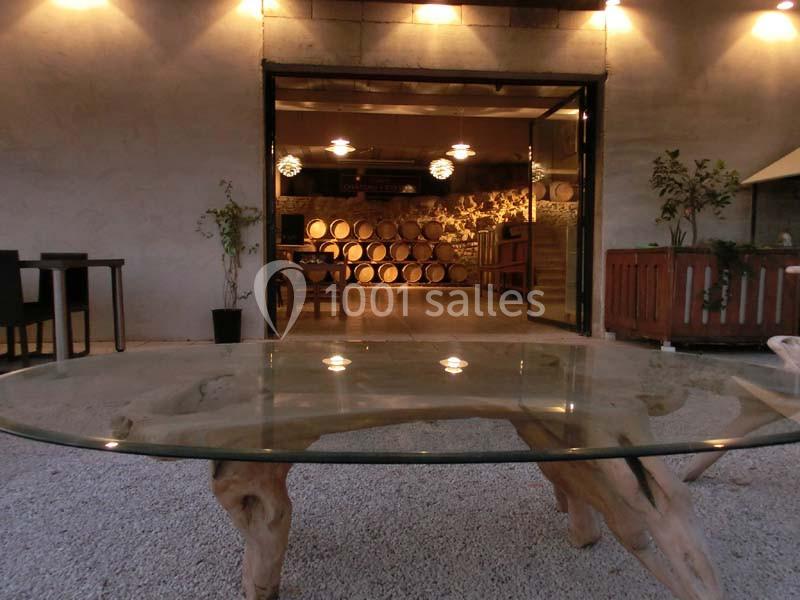 Salle de dégustation avec des tonneaux en arrière-plan, vue depuis une table en verre sur une terrasse en gravier.