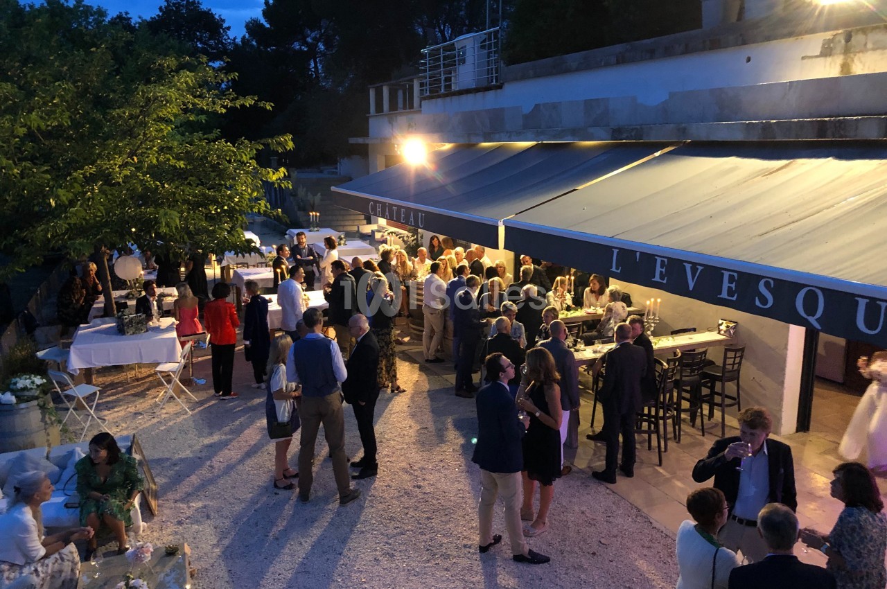 Groupe de personnes rassemblées en soirée près d'une terrasse éclairée, avec tables et arbres en arrière-plan.