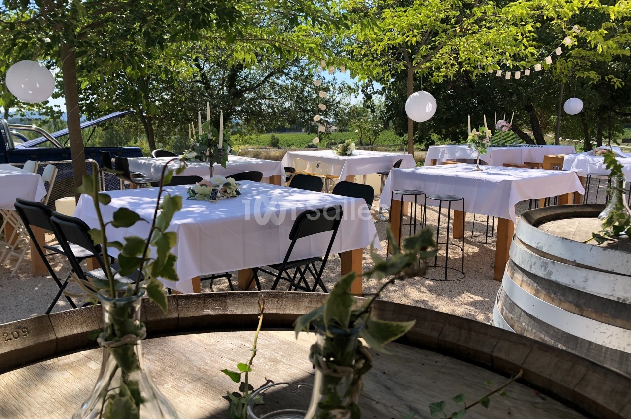 Tables décorées avec nappes blanches et bouquets, disposées en extérieur sous des arbres pour un événement festif.