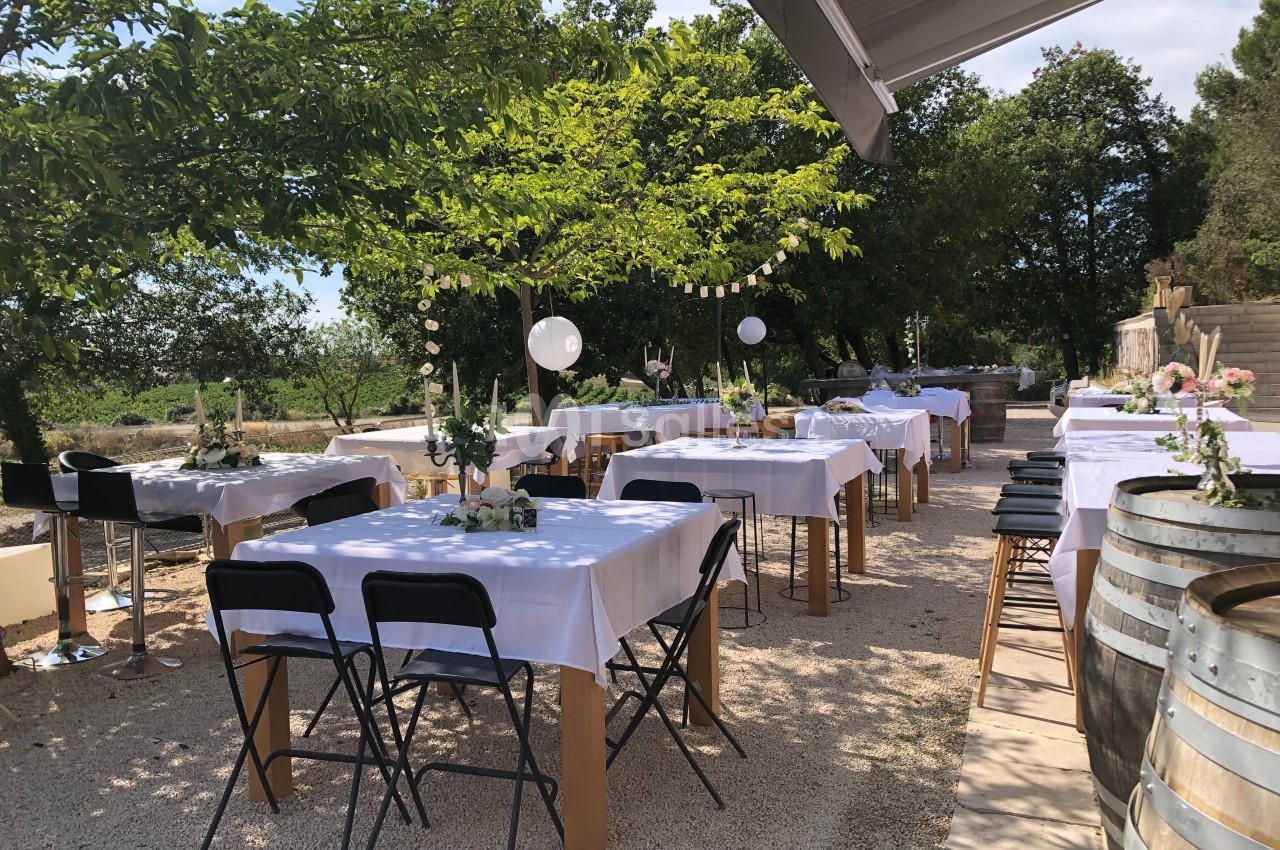 Tables dressées avec nappes blanches sous des arbres, décorées pour un événement en extérieur par temps ensoleillé.