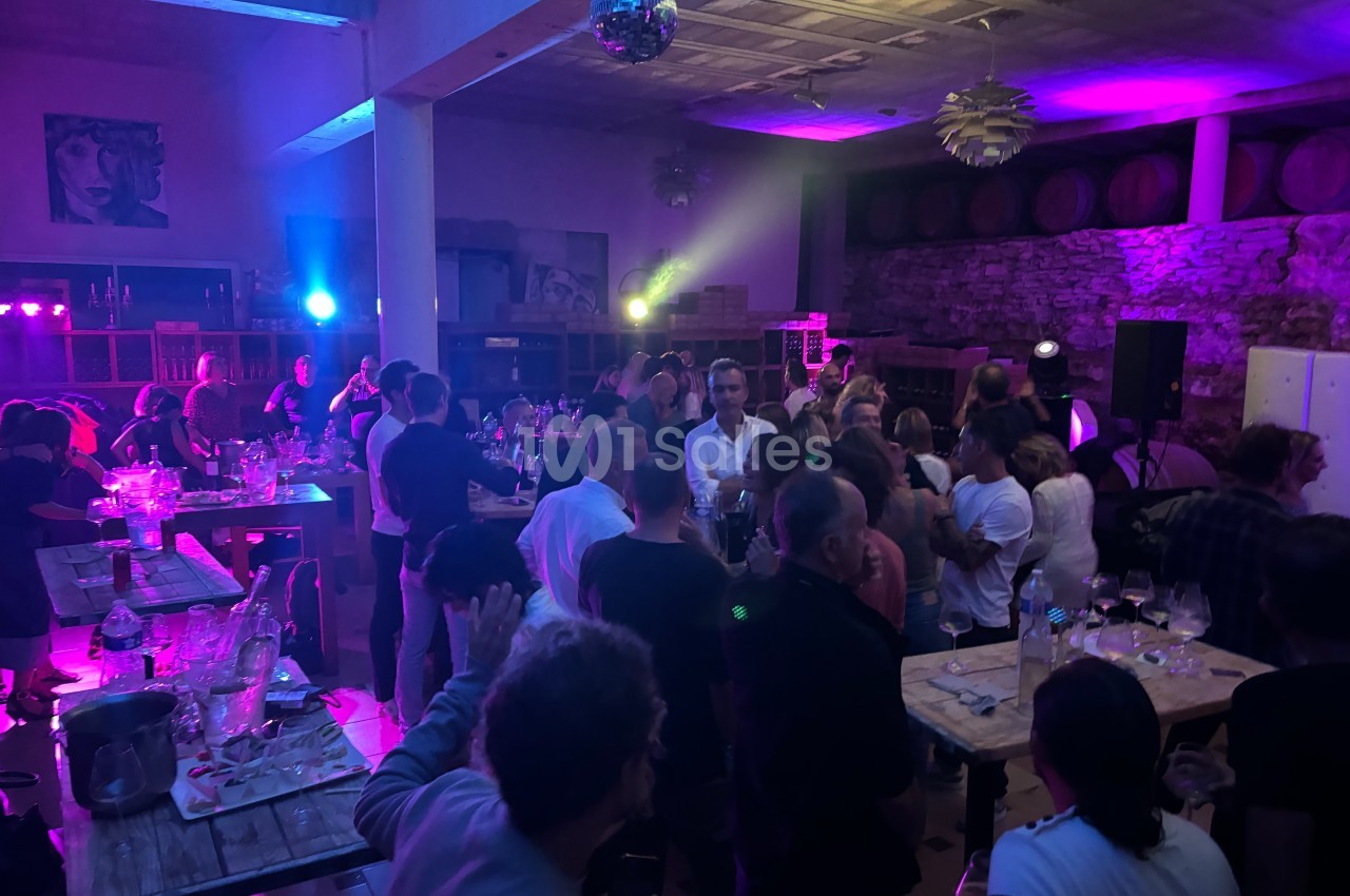 Groupe de personnes rassemblées dans une salle éclairée par des lumières colorées, avec tables et tonneaux en arrière-plan.