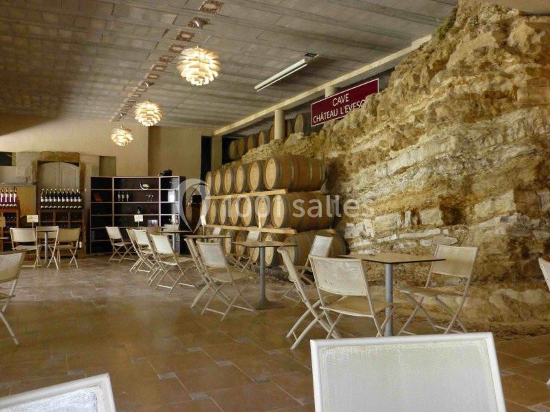 Salle de dégustation avec tables, chaises, murs en pierre et rangées de fûts de bois alignés.