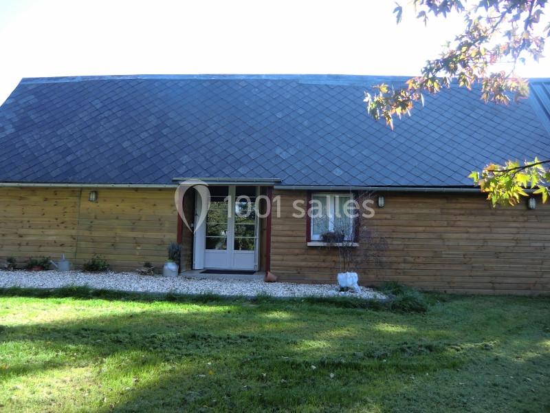 Façade d'une maison en bois avec toit en ardoise, porte vitrée centrale et petite fenêtre à droite, entourée de pelouse.