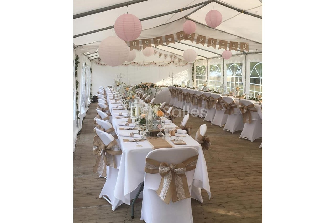 Salle de réception décorée pour un mariage, avec tables alignées, nappes blanches et chaises ornées de rubans beige.