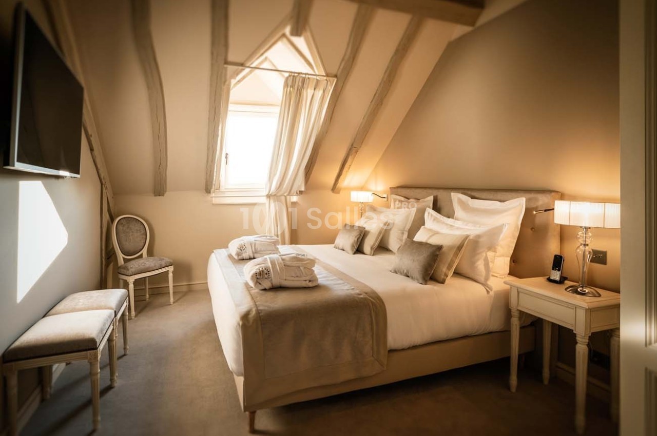 Chambre mansardée avec lit double, poutres apparentes, éclairage doux, télé murale et mobilier élégant.