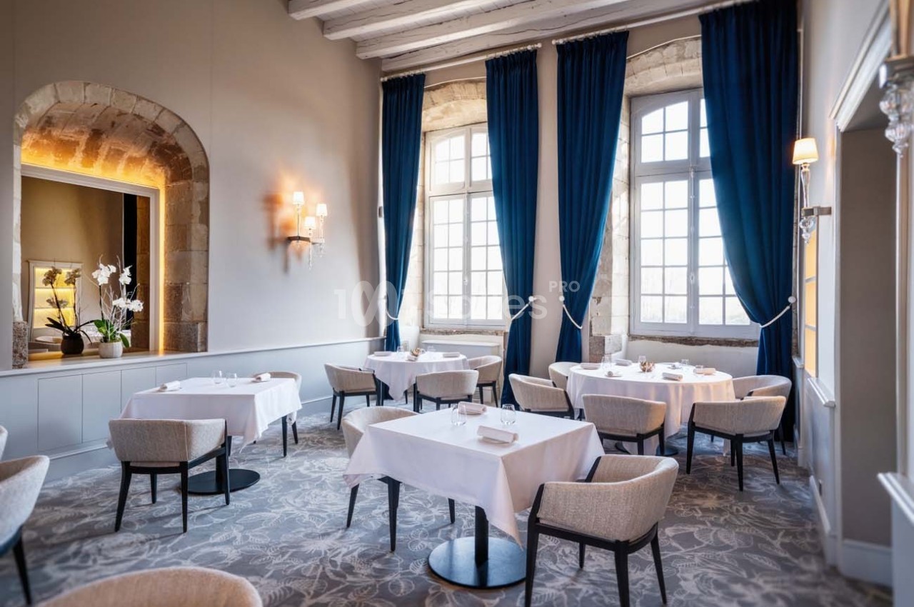 Salle de restaurant élégante avec tables dressées, grandes fenêtres encadrées de rideaux bleus et murs en pierre apparente.