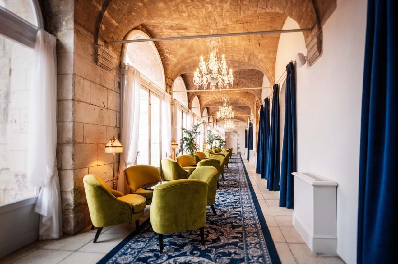 Couloir lumineux avec fauteuils verts, tapis bleu, rideaux foncés et lustres suspendus sous des voûtes en pierre.