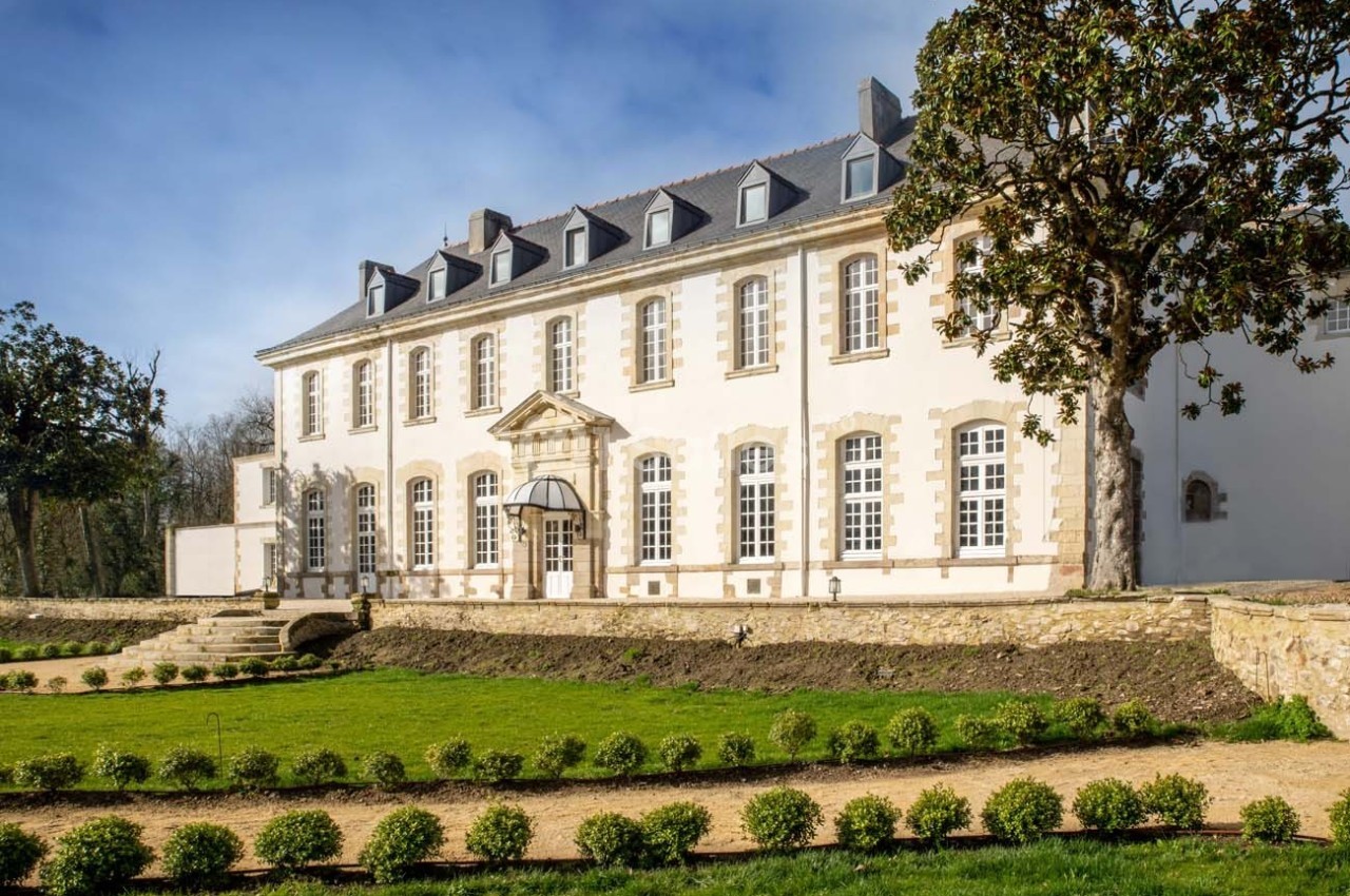 Façade d'un grand bâtiment historique en pierre, entouré d'un jardin paysager avec pelouse et arbres.