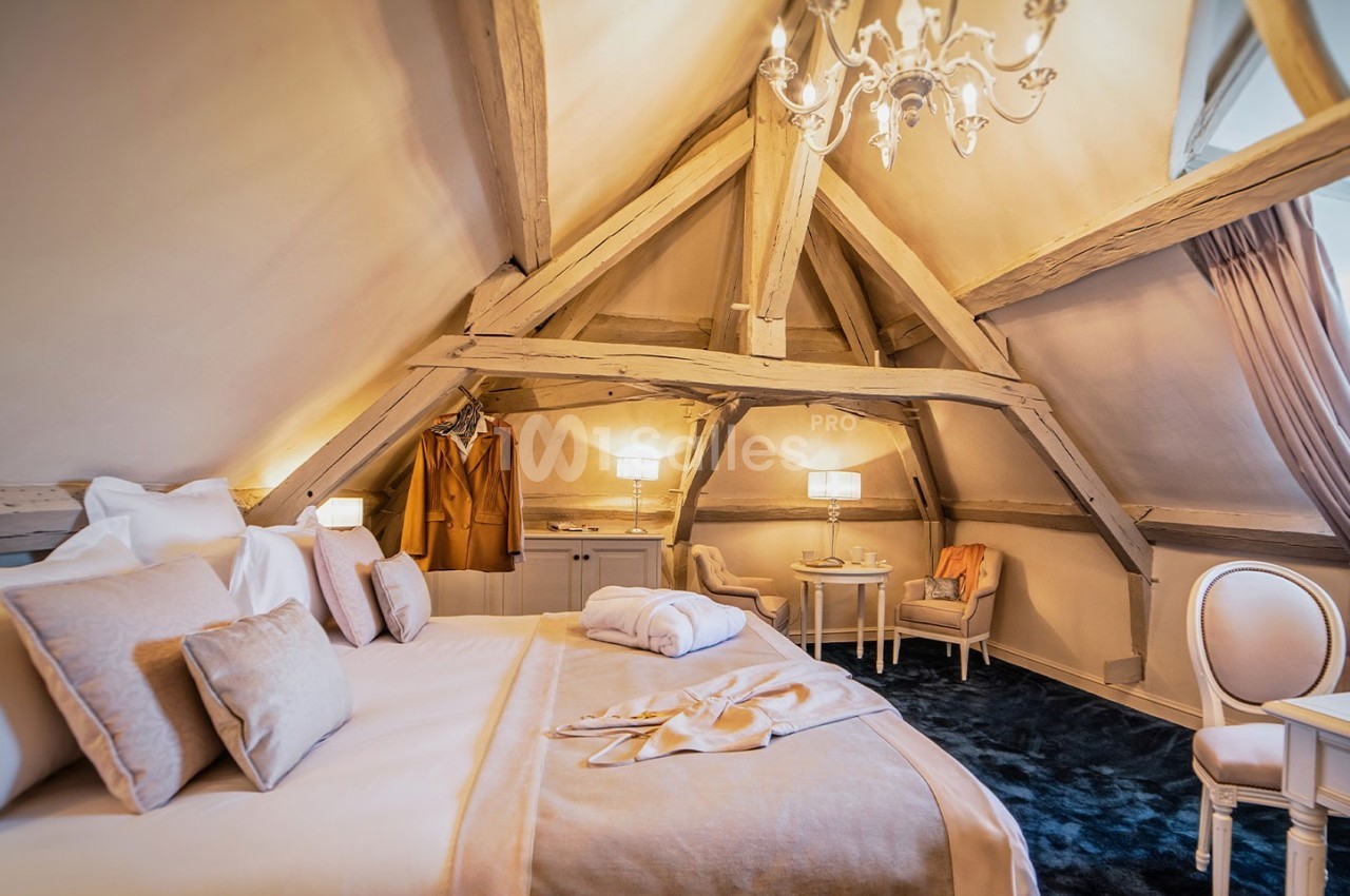 Chambre mansardée avec poutres apparentes, lit double, fauteuils, table et éclairage chaleureux sous un lustre.