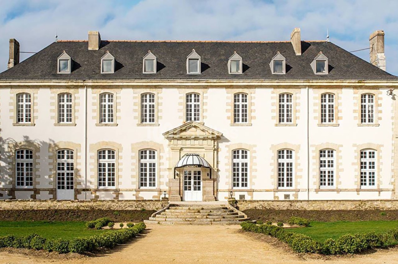 Façade d'un bâtiment ancien en pierre avec des fenêtres symétriques et un toit en ardoise sous un ciel dégagé.