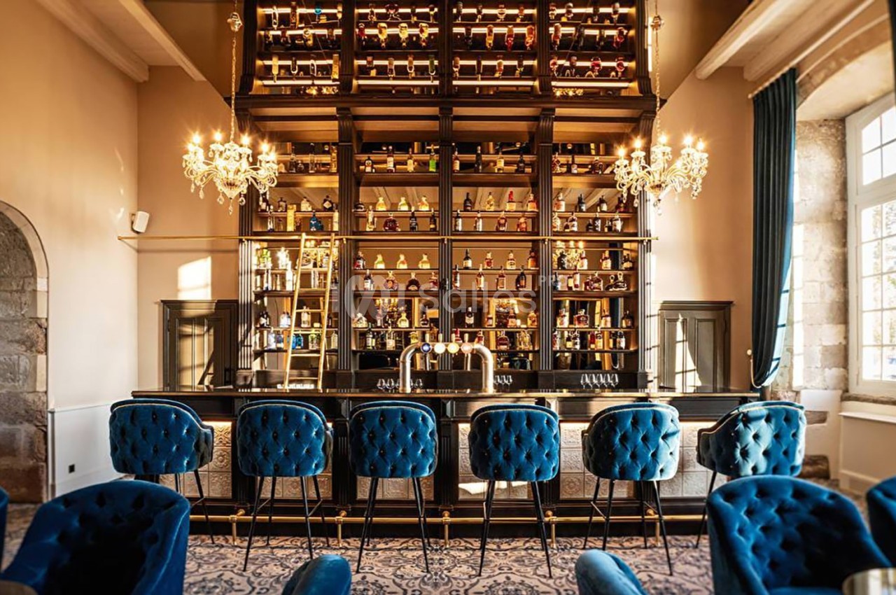 Bar élégant avec des tabourets en velours bleu, étagères remplies de bouteilles et lustres suspendus dans une ambiance…