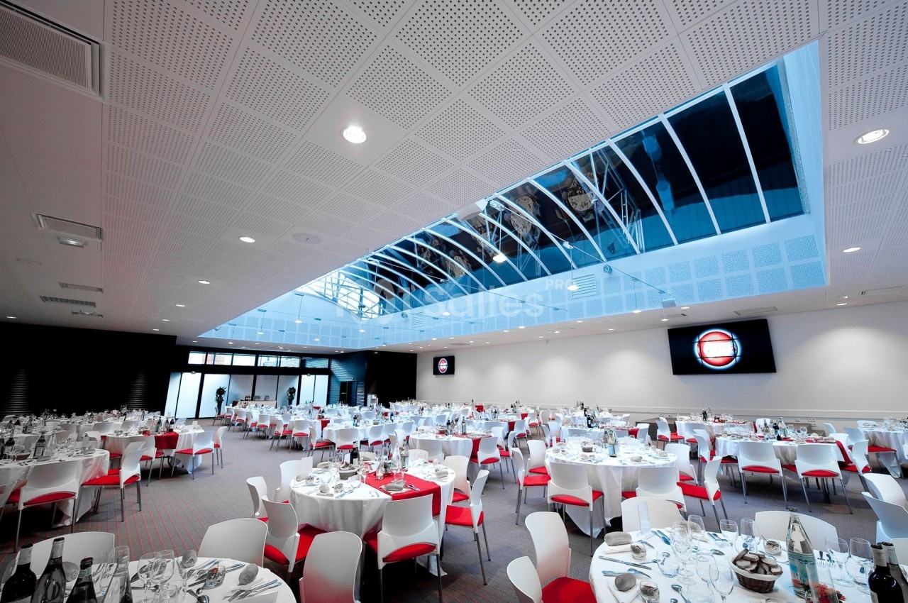 Salle de réception lumineuse avec tables rondes dressées, chaises blanches et rouges, et plafond vitré.