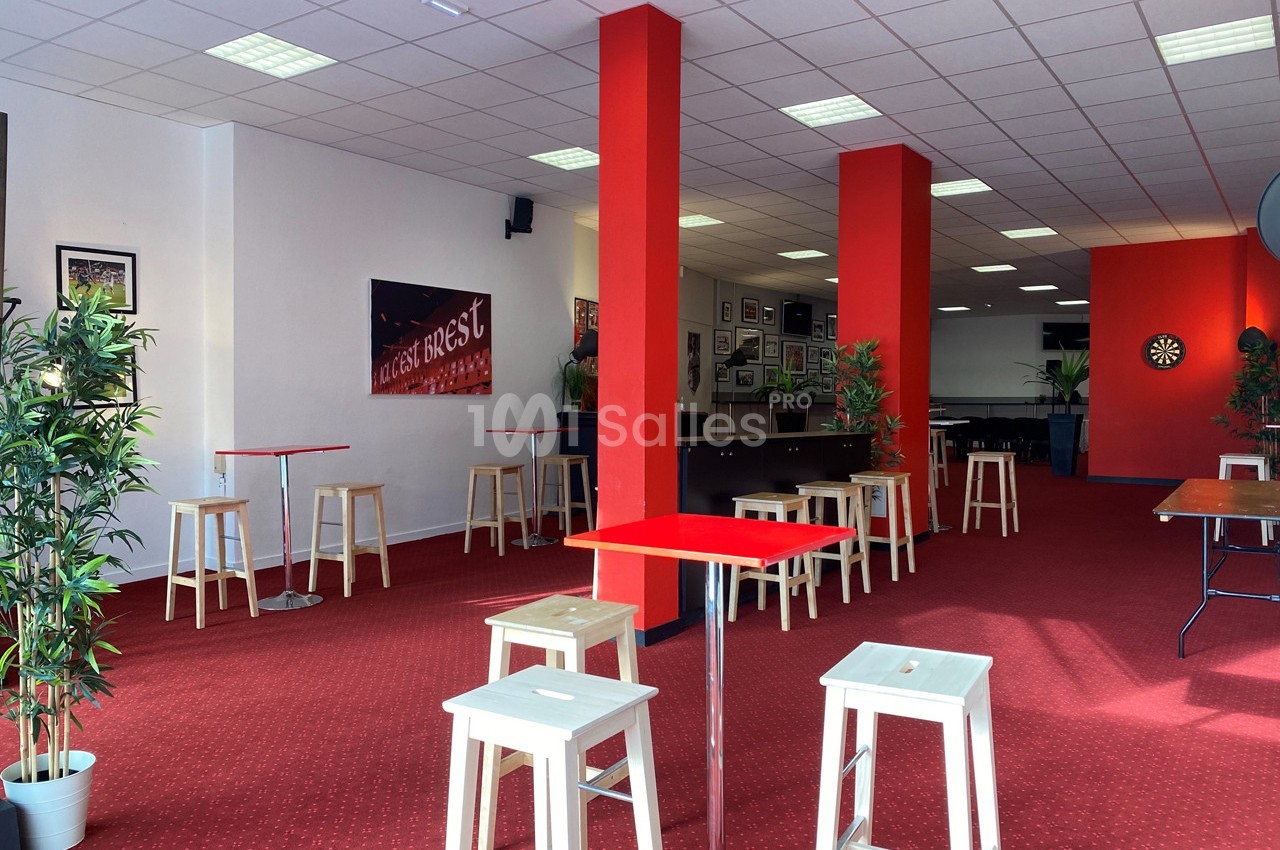 Salle lumineuse avec moquette rouge, tables hautes, tabourets en bois, plantes en pot et cible de fléchettes au fond.