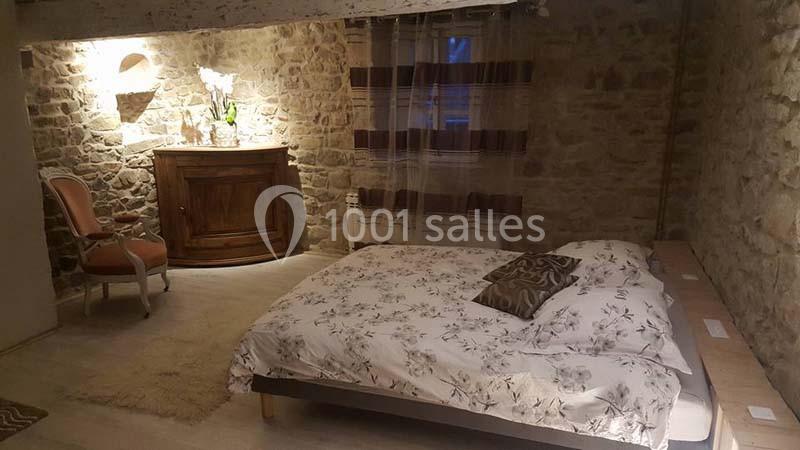 Chambre avec murs en pierre apparente, lit double avec housse florale, fauteuil et meuble en bois sous éclairage doux.