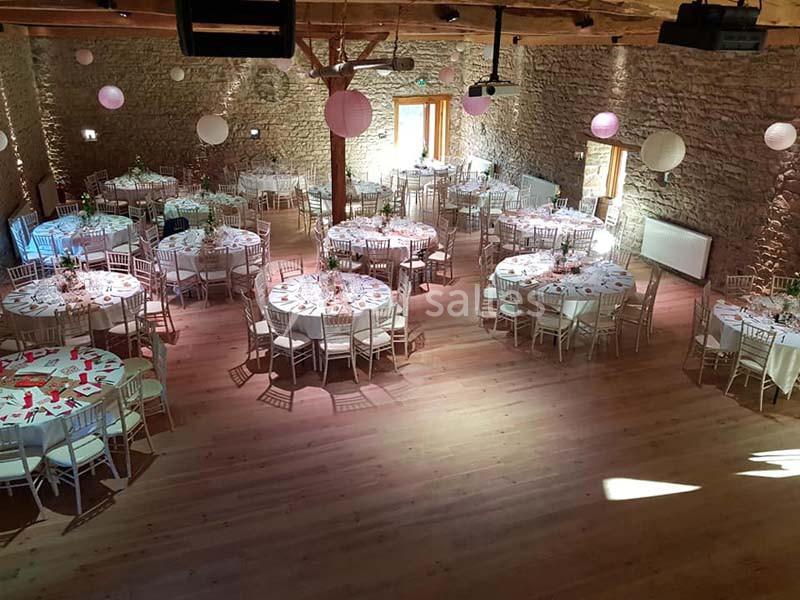 Salle de réception en pierre avec tables rondes dressées pour un événement, éclairée par la lumière naturelle.