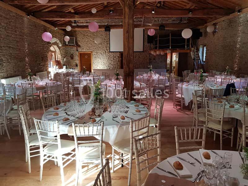 Salle de réception en pierre avec tables rondes dressées, chaises en bois clair et décorations suspendues.