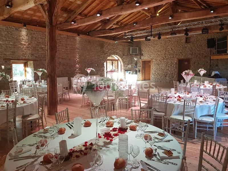 Salle de réception en pierre avec tables rondes dressées pour un événement, décorées de fleurs et pétales rouges.