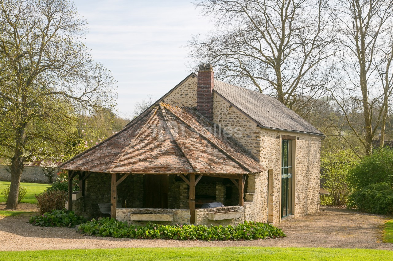 Bâtiment en pierre avec toit en tuiles et auvent en bois, entouré de verdure et d'arbres en arrière-plan.