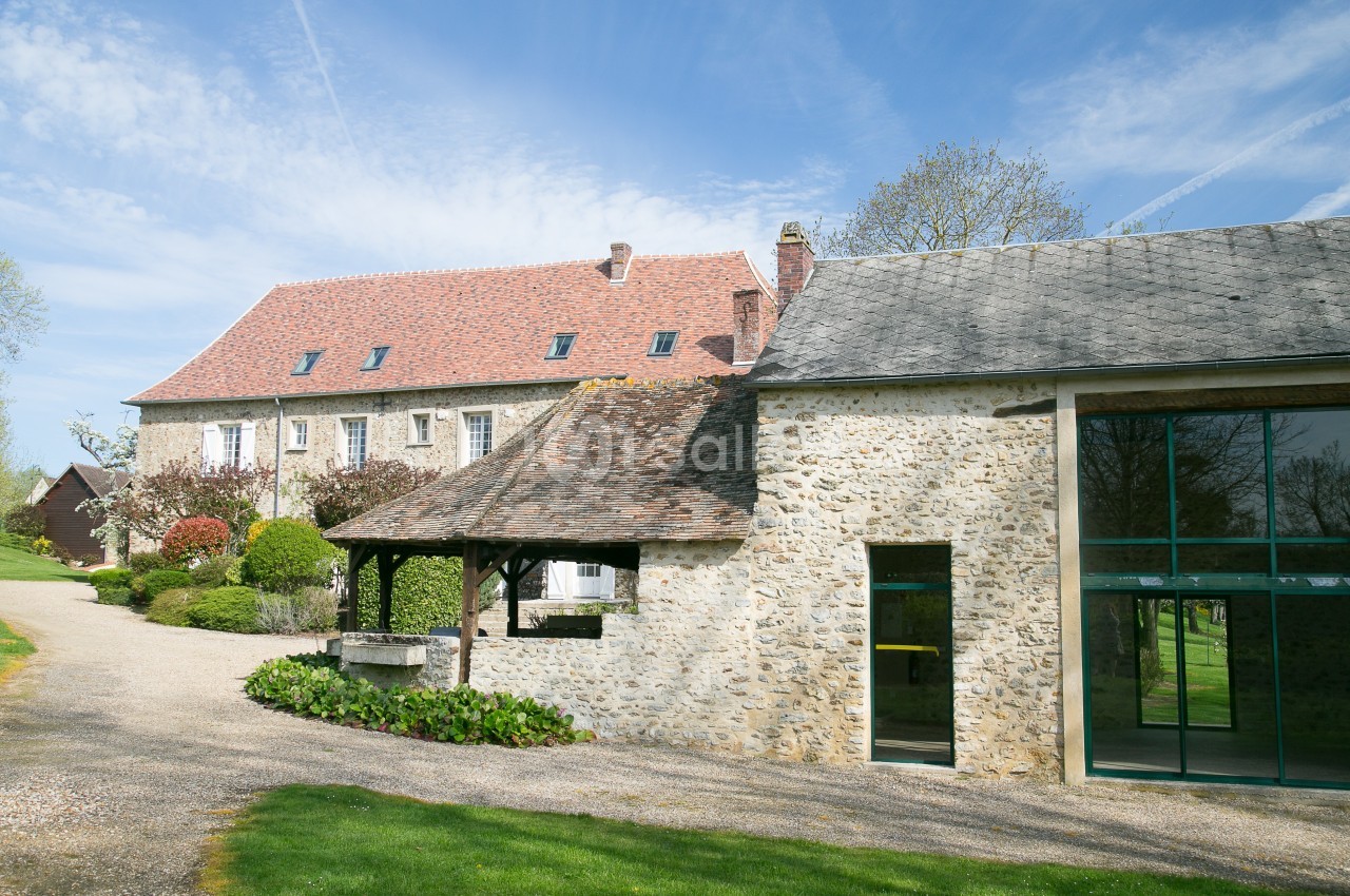 Bâtiment en pierre avec toit en tuiles, grandes fenêtres modernes et jardin verdoyant sous un ciel bleu.