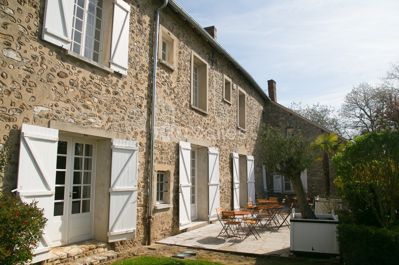 Façade en pierre d'une maison avec volets blancs, terrasse aménagée et jardin arboré sous un ciel clair.