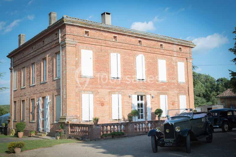 Façade d'une maison en briques rouges avec volets blancs, une voiture ancienne stationnée devant.