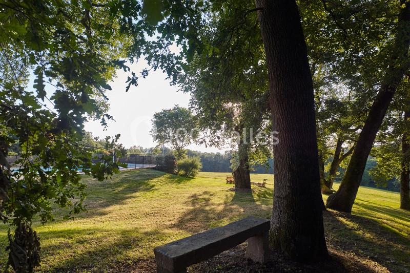 Vue d'un parc arboré avec une clairière ensoleillée, des bancs en bois et une piscine visible à l'arrière-plan.