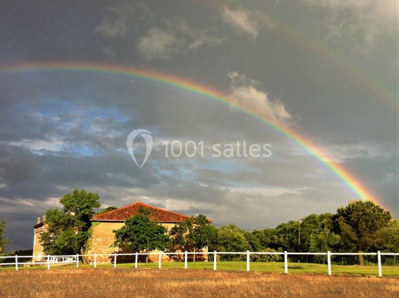 Un double arc-en-ciel au-dessus d'une maison en pierre entourée de verdure et d'une clôture blanche.