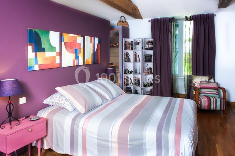 Chambre lumineuse avec murs violets, lit à rayures pastel, tableaux colorés et étagères décoratives près d'une fenêtre.