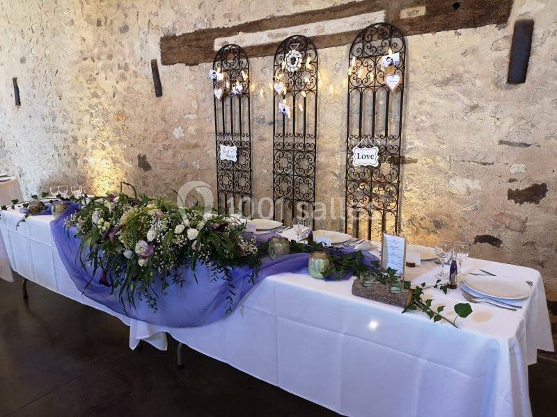 Table de réception décorée avec des fleurs, un chemin de table violet et des éléments rustiques sur fond de mur en pierre.