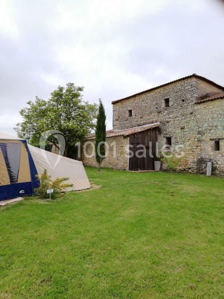 Tente beige et bleue installée sur une pelouse verte près d'un bâtiment en pierre avec un arbre et un ciel nuageux.