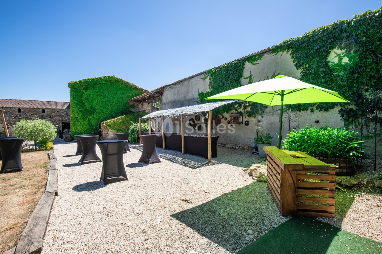 Cour extérieure avec tables hautes noires, parasol vert, végétation et murs en pierre recouverts de lierre.