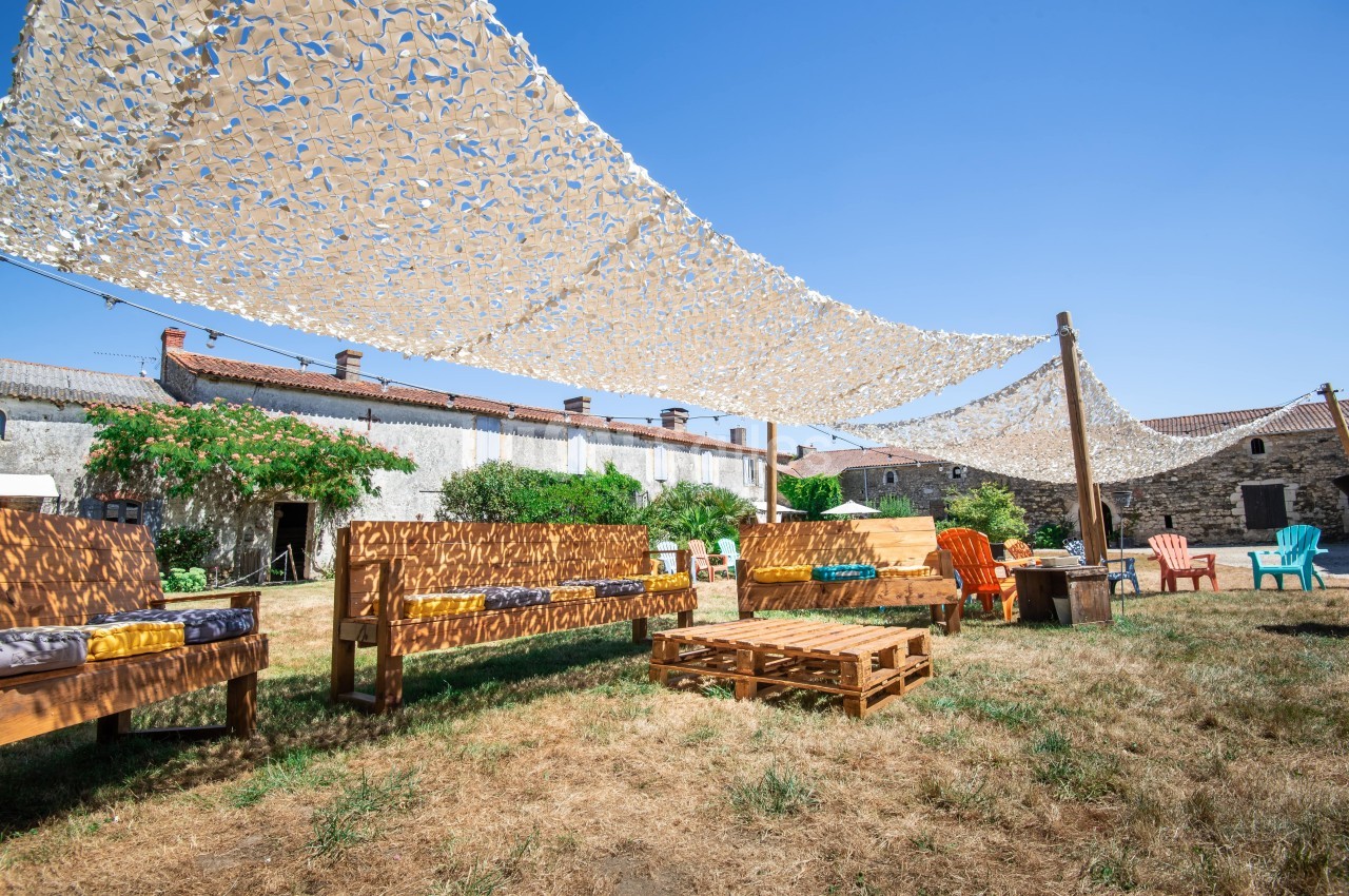 Espace extérieur avec bancs en bois, palettes et chaises sous des voiles d'ombrage, entouré de bâtiments en pierre.