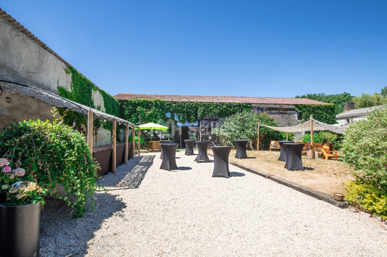 Cour extérieure avec gravier, tables hautes noires, parasols, végétation et bâtiments en pierre sous un ciel bleu.