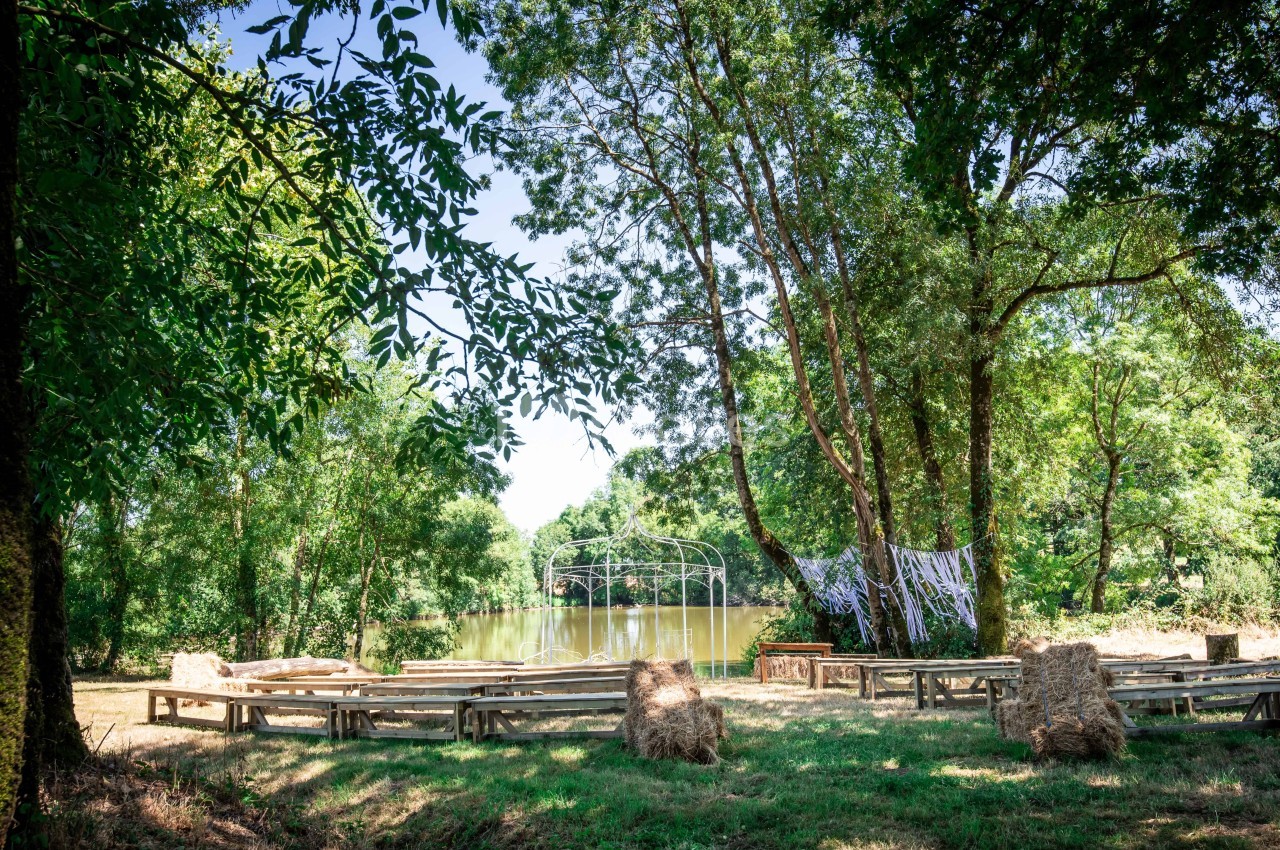 Espace extérieur avec bancs en bois et bottes de paille, face à un lac entouré d'arbres sous un ciel dégagé.