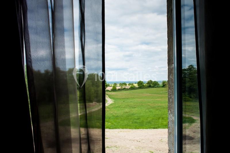 Vue sur un paysage champêtre verdoyant à travers une fenêtre partiellement couverte de rideaux sombres.