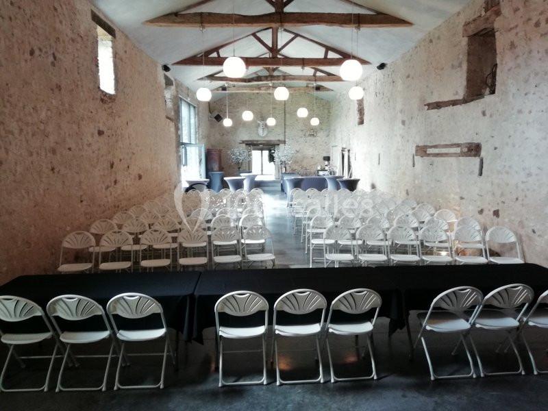 Salle en pierre avec des rangées de chaises blanches alignées face à une table noire, éclairée par des suspensions rondes.