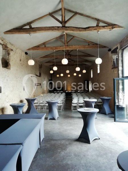 Salle en pierre avec poutres apparentes, aménagée pour un événement avec chaises alignées et tables hautes drapées.