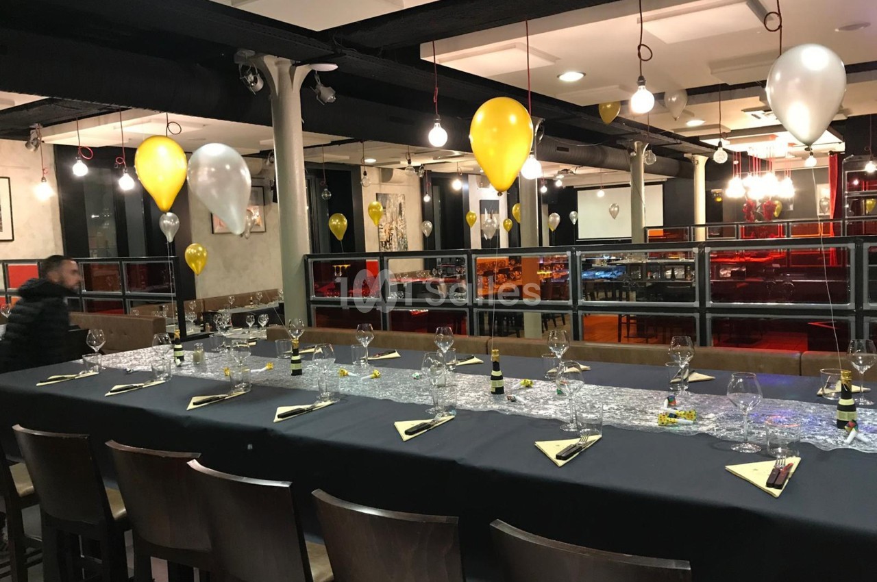 Grande table dressée avec nappes noires, verres et décorations, entourée de chaises dans une salle éclairée avec ballons…
