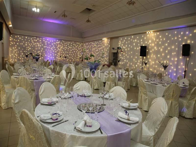 Salle de réception décorée avec des tables rondes, nappes blanches, chaises couvertes et guirlandes lumineuses au mur.