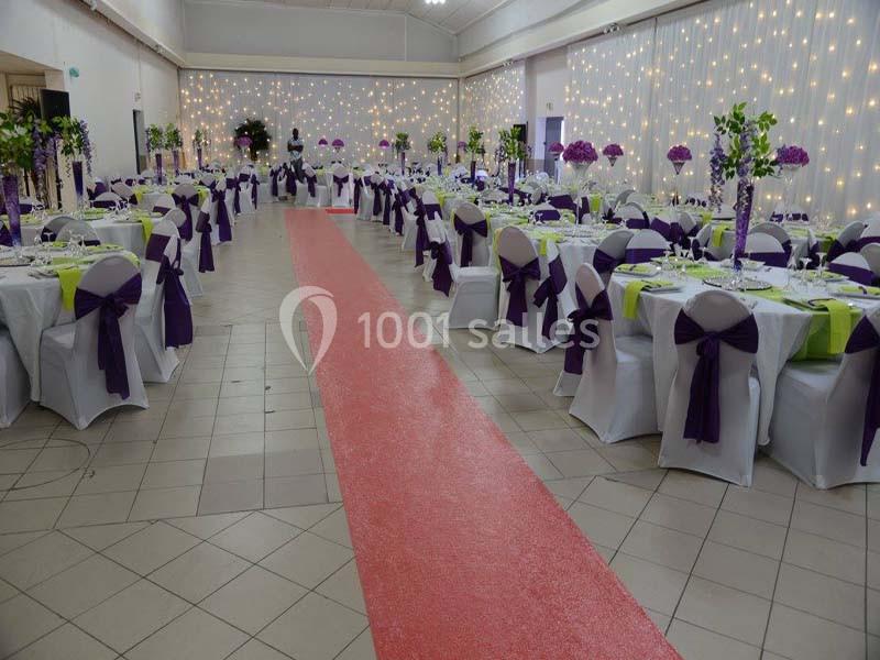 Salle de réception décorée avec des tables dressées, nappes blanches, rubans violets et guirlandes lumineuses en arrière…