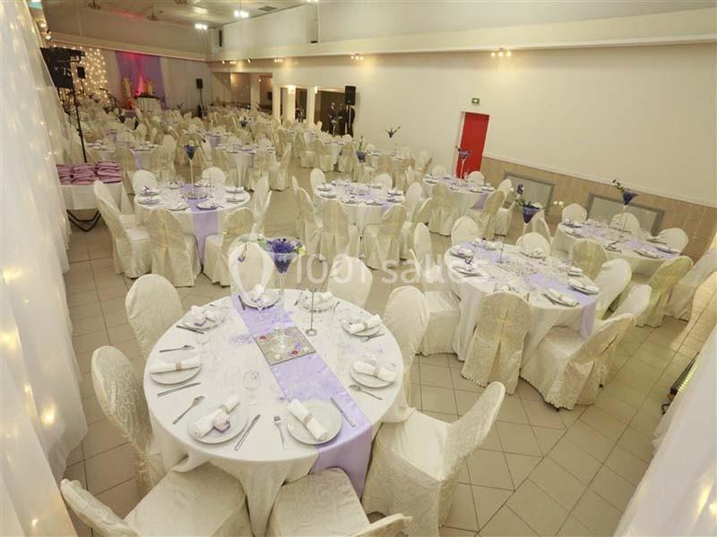 Salle de réception décorée avec des tables rondes, nappes blanches, chemins de table mauves et chaises couvertes.