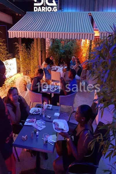 Des personnes dînent en terrasse le soir, sous des auvents rayés, entourées de bambous et d'une lumière chaleureuse.