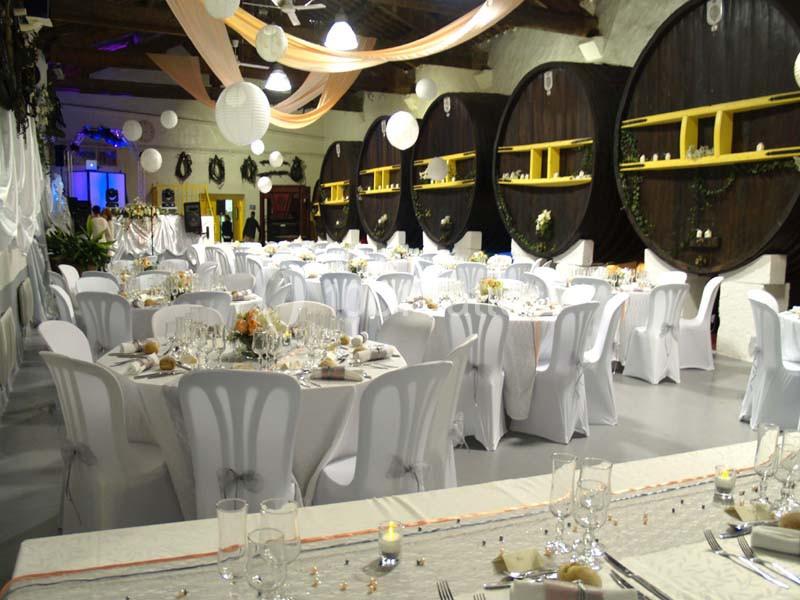 Salle de réception décorée avec des tables rondes dressées, des chaises blanches et des tonneaux en arrière-plan.