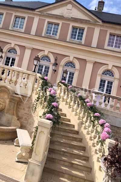 Escalier en pierre décoré de fleurs roses, menant à une façade classique avec fenêtres cintrées et balustrades.