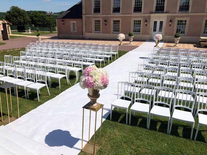 Chaises blanches alignées en extérieur pour une cérémonie, avec une allée centrale et des arrangements floraux.